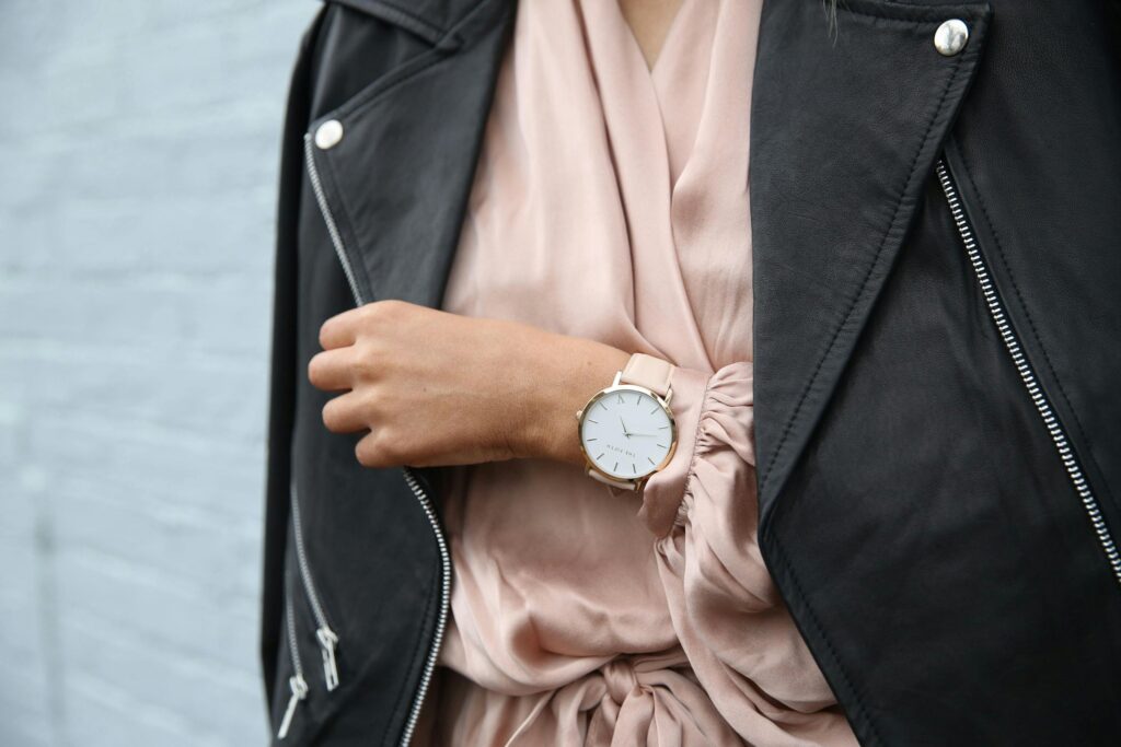 Femme portant un gilet noir, une robe rose et une montre dorée. Il y a un mur gris derrière elle.